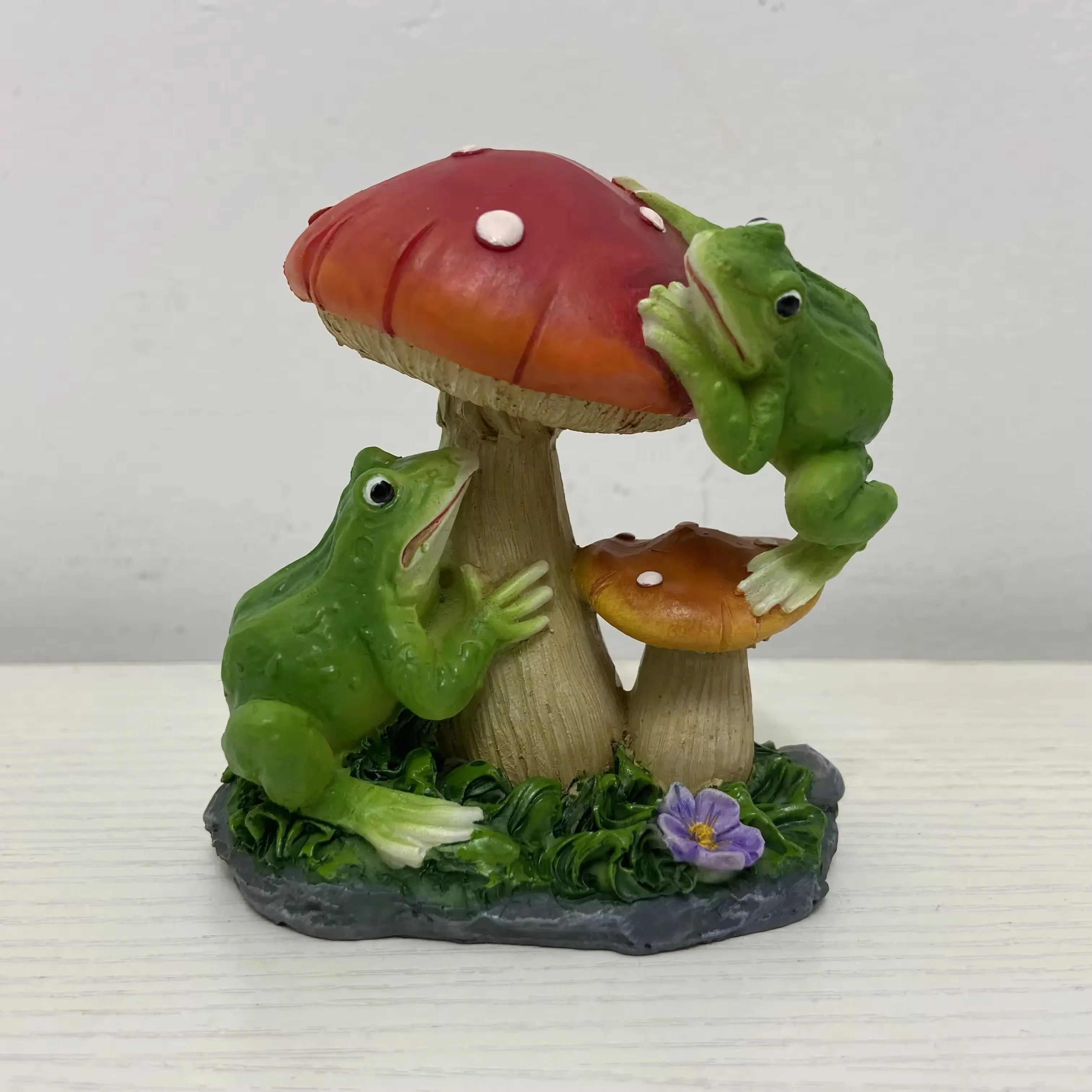 Two resin frog figurines climbing around red and orange mushrooms in a playful garden ornament scene