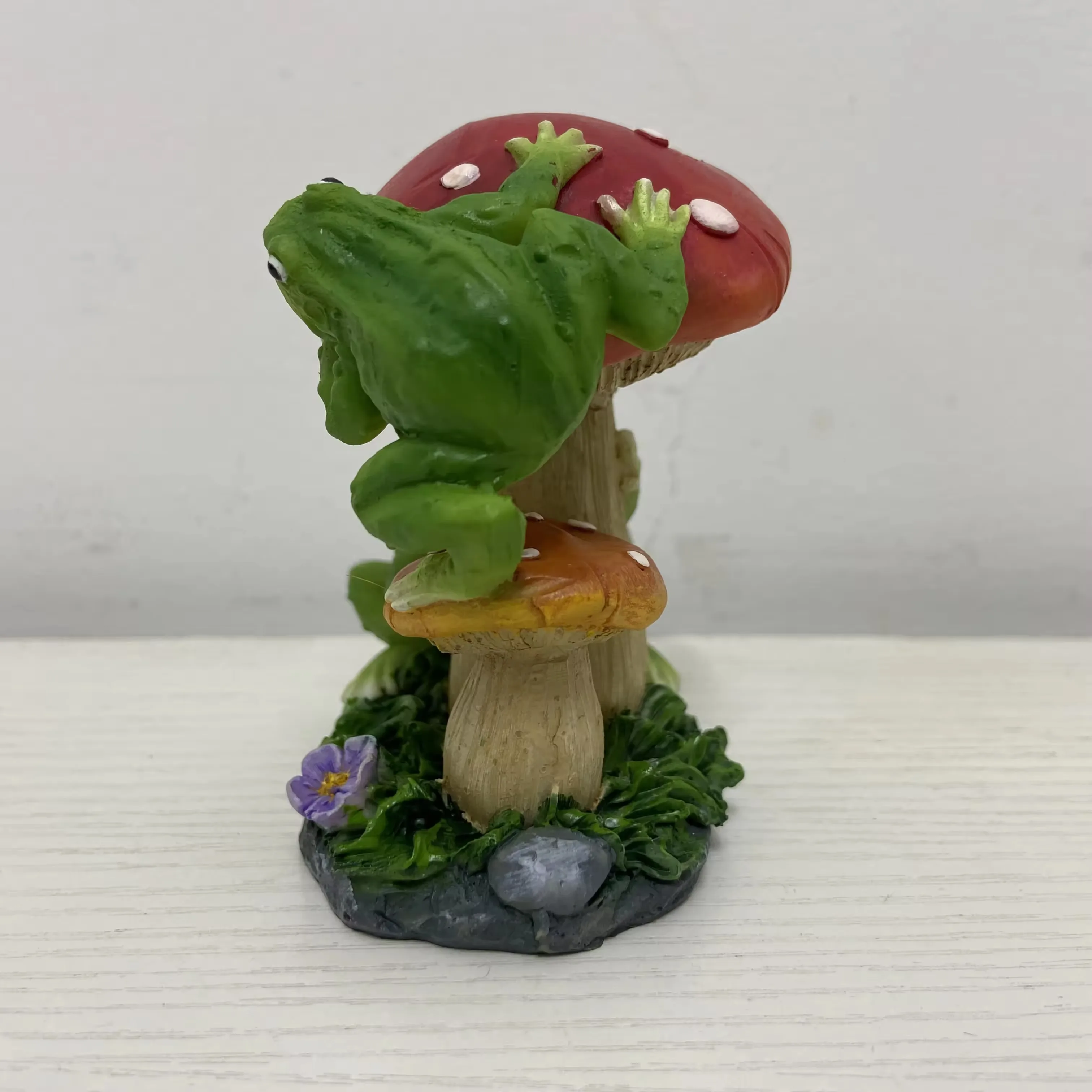 Rear view of a hand-painted resin frog climbing a mushroom garden ornament with floral base detail