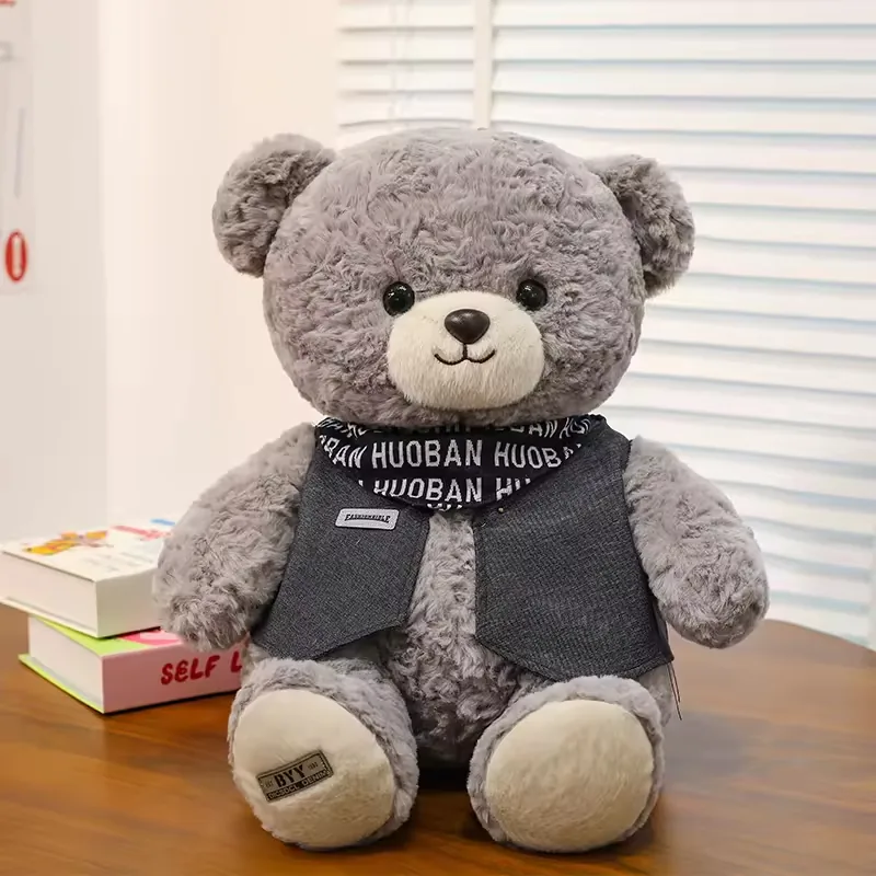Grey teddy bear plush doll seated upright with curly fur, cream muzzle, black scarf, and dark grey vest